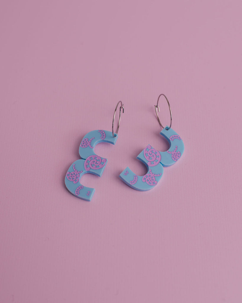 MISS MAIA, May 2022. A flatlay pic of the Blue and Lilac Kōtuitui I earrings on a pink background.